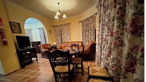 A dining area with a brown leather table and chairs, a television on a stand, and floral curtains. - Turismo rural Valsequillo (Valsequillo)