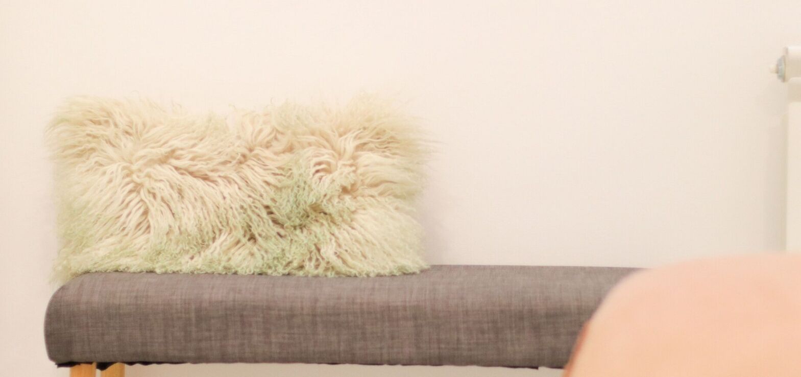 A grey upholstered bench with a fluffy beige pillow against a plain wall.