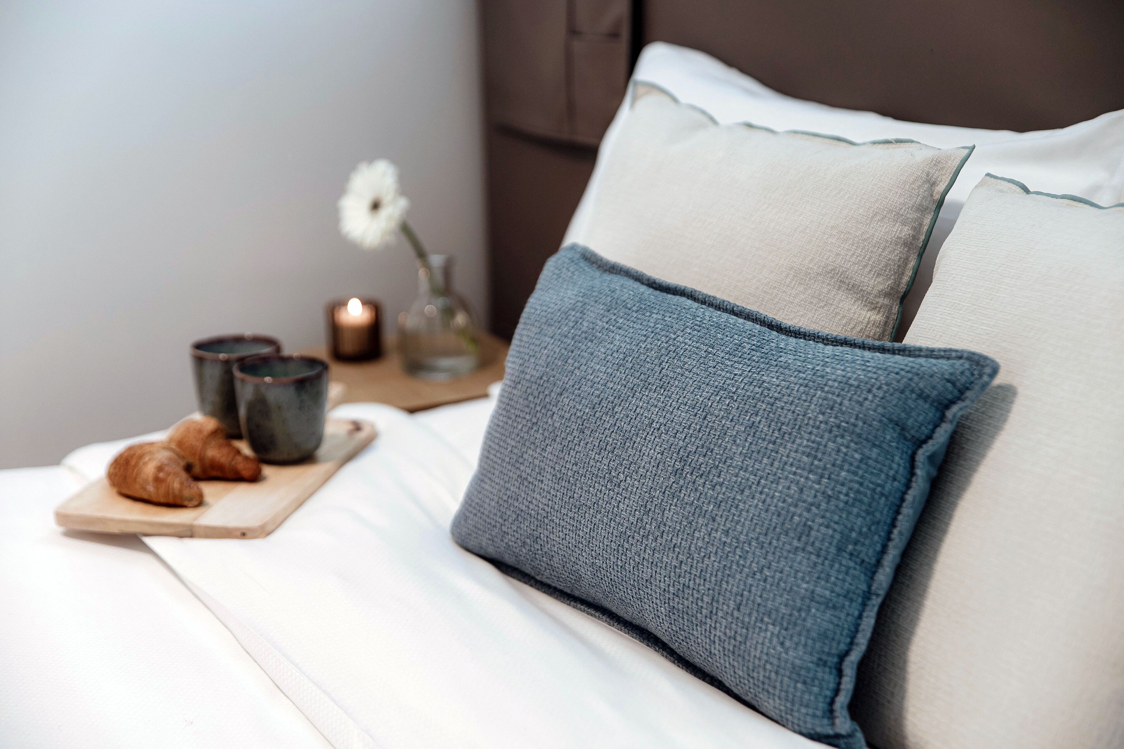 Un lit bien fait, avec un oreiller bleu et un oreiller beige, un plateau en bois avec des croissants, une bougie et un vase avec une fleur.