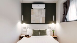 Ein ordentlich bezogenes Bett mit weißer Decke und zwei Kissen, ein Nachttisch mit Buch und Tasse sowie eine Wand-Klimaanlage über dem Bett.
