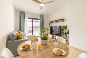 A modern living room with a dining table set for breakfast, a sofa, a TV, and a view of the outdoors.