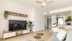 A modern living room with a flat-screen TV, a ceiling fan, a sofa, a dining area, and a kitchen in the background.