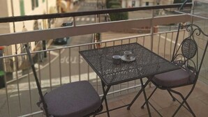 A balcony with a metal table and two chairs, overlooking a coastal town with buildings and a hilly landscape.