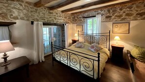 A bedroom with a stone wall, a bed with a metal frame, two bedside tables with lamps, and a window with curtains. - La Vigne: modernized farmhouse from 1882, incl. swimming pool (from August 2025) (Saint-Flour-l'Étang)