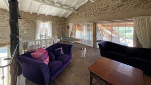 A spacious room with stone walls, a high ceiling with exposed beams, a wooden table, and a purple sofa. - La Vigne: modernized farmhouse from 1882, incl. swimming pool (from August 2025) (Saint-Flour-l'Étang)