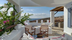 Une terrasse aux murs blancs, un canapé en osier et une table agrémentée de coussins et d’un panier de fruits.