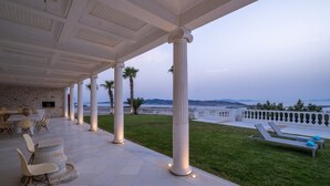 Un'area esterna coperta con pavimento in pietra, colonne bianche e vista sul mare.