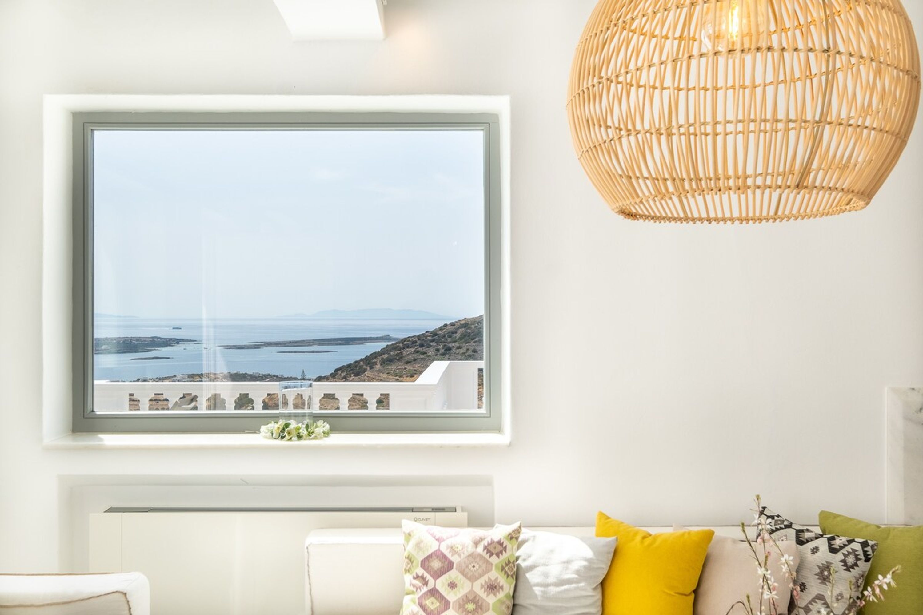 Ein Zimmer mit großem Fenster und Blick auf eine Küstenlandschaft, ein weißes Sofa mit bunten Kissen sowie eine gewebte Pendelleuchte.