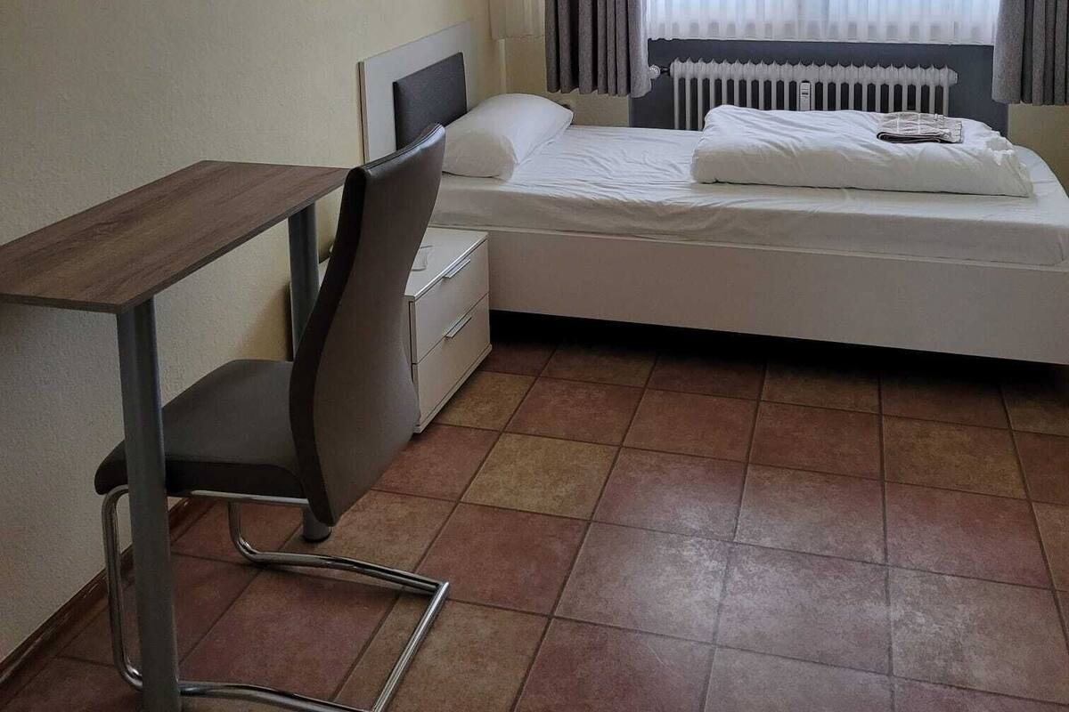 A hotel room with a bed, a desk, a chair, a radiator, and a window with curtains.