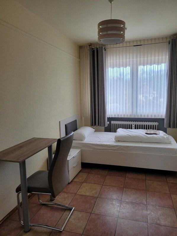 A hotel room with a bed, a desk, a chair, a radiator, and a window with curtains. - Horrem City Apartments (Kerpen)