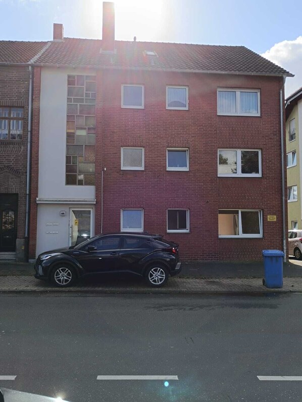A red brick building with multiple windows, a car parked in front, and a blue trash bin. - Horrem City Apartments (Kerpen)