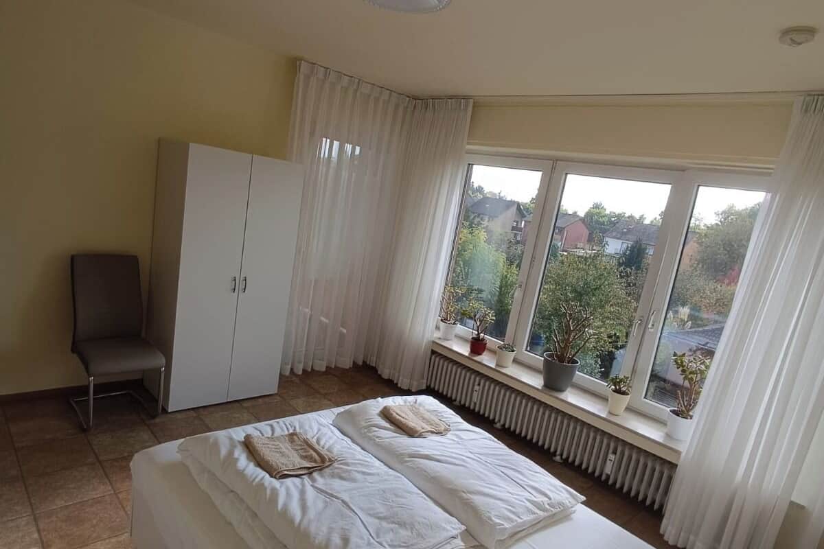 A bedroom with a large bed, a chair, a wardrobe, and a window with a view of the outside.