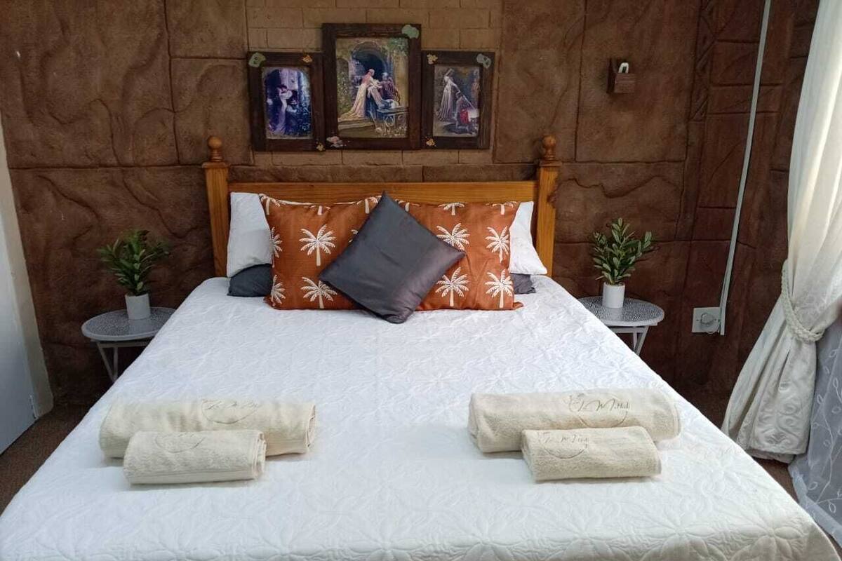 A neatly made bed with white linens and a few decorative pillows, two small potted plants on side tables, and framed artwork on the wall.