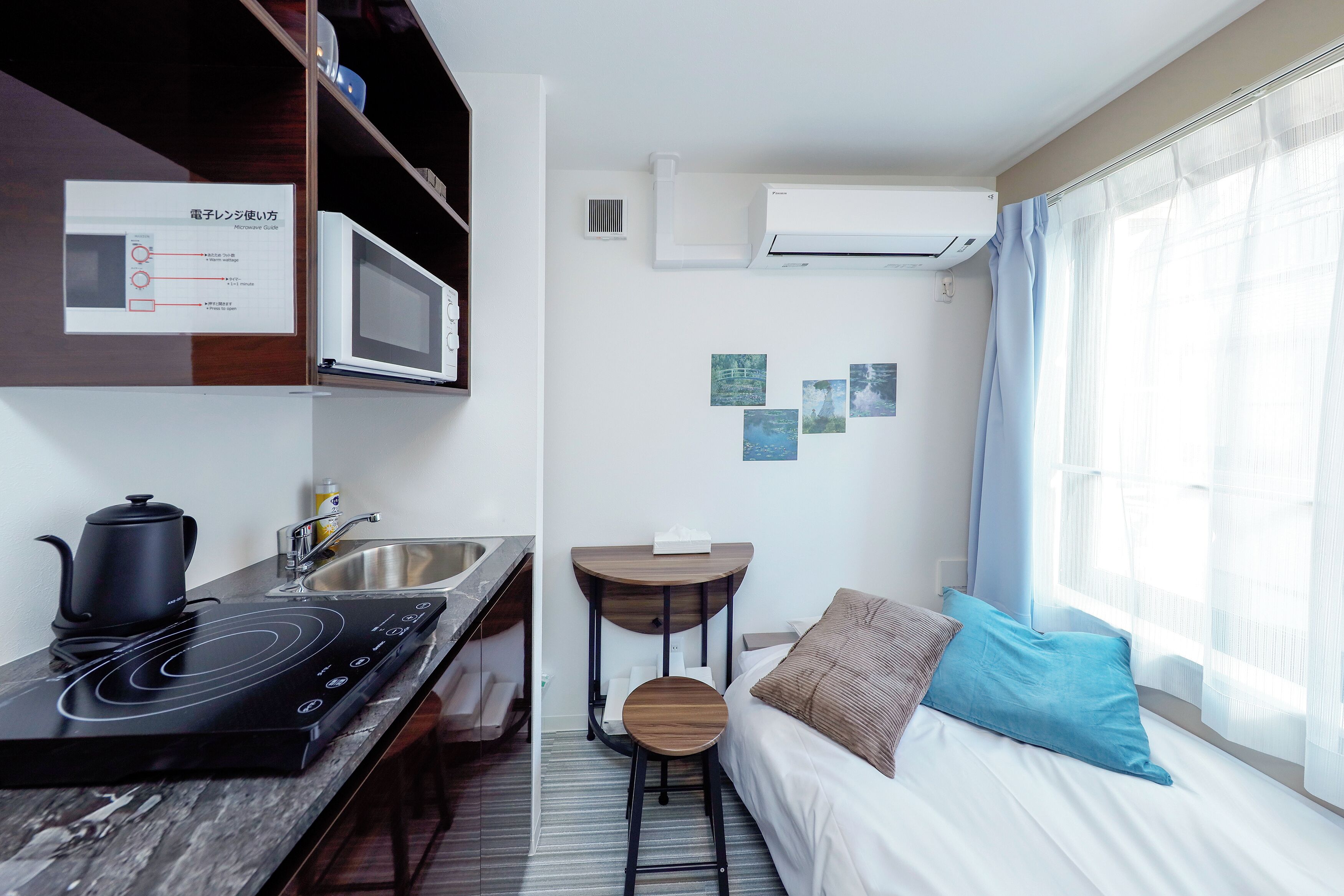 A compact hotel room with a kitchenette, a bed with a blue pillow, and a window with sheer curtains.