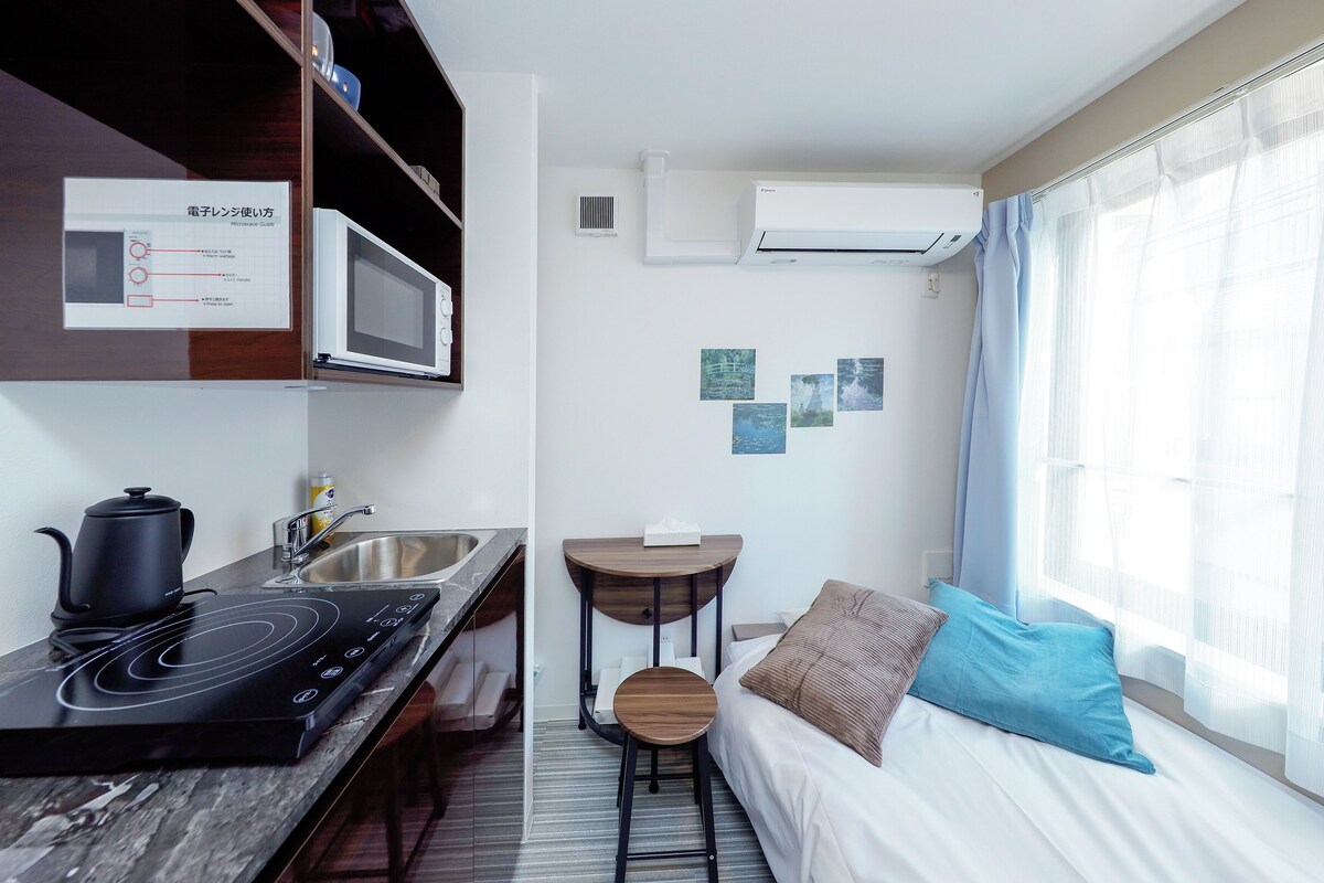 Una camera d'albergo compatta con angolo cottura, un letto con cuscino blu e una finestra con tende trasparenti.