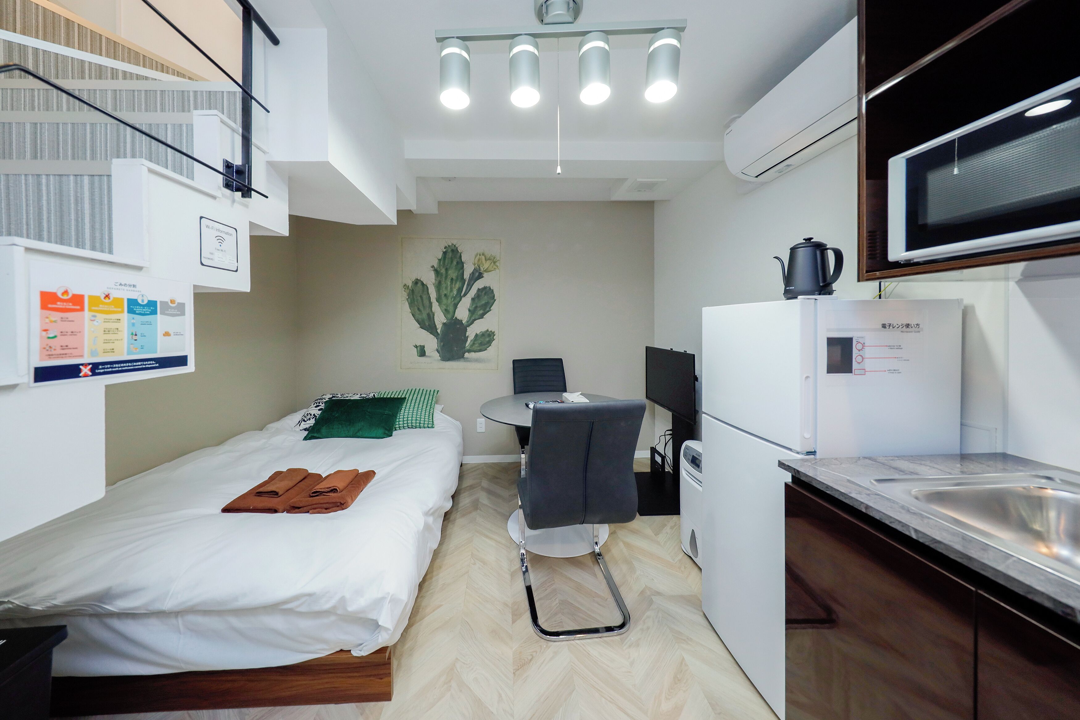 Habitación de hotel pequeña con cama, escritorio, silla, microondas, lavamanos y lámpara en el techo.