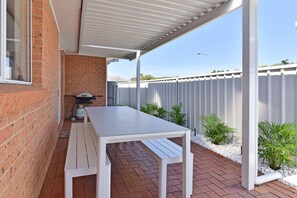 Une terrasse avec une table et des bancs blancs, un barbecue et un mur en briques.