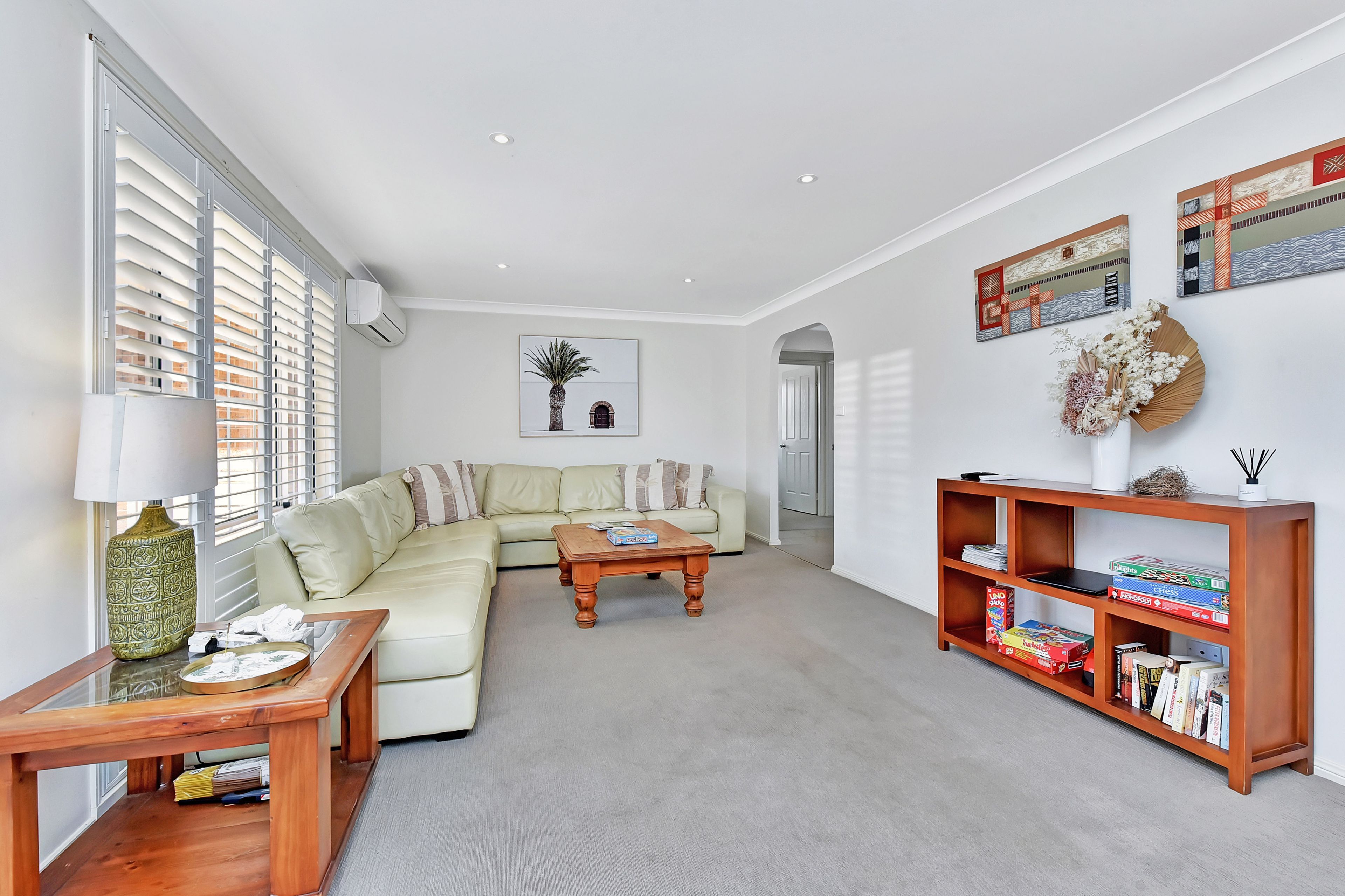 A modern living room with a sectional sofa, a wooden coffee table, a bookshelf, and a large window with blinds.