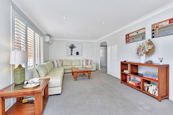A modern living room with a sectional sofa, a wooden coffee table, a bookshelf, and a large window with blinds.