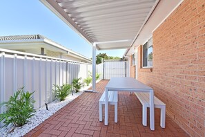 Un patio couvert avec une table et des bancs blancs, entouré d’un mur de briques et d’une clôture blanche.