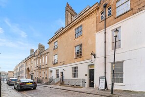 A row of traditional buildings with a cobblestone street, a black car parked on the side, and a signboard on the right. - Experience Elegance in Central Bath (Bath)