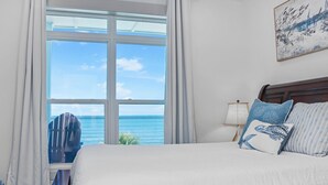 A bedroom with a large window offering a view of the ocean, a bed with blue and white pillows, and a framed artwork on the wall. - Just Beachy (Port St. Joe)