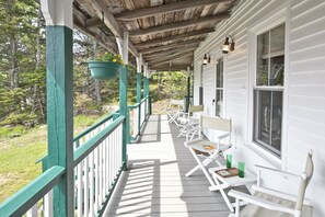 Une véranda couverte avec des balustrades blanches et un plafond en bois, comprenant un espace salon extérieur et des jardinières suspendues.