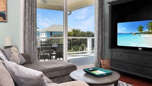 A living room with a large TV displaying a tropical beach scene, a sofa, a coffee table, and a balcony with outdoor furniture.