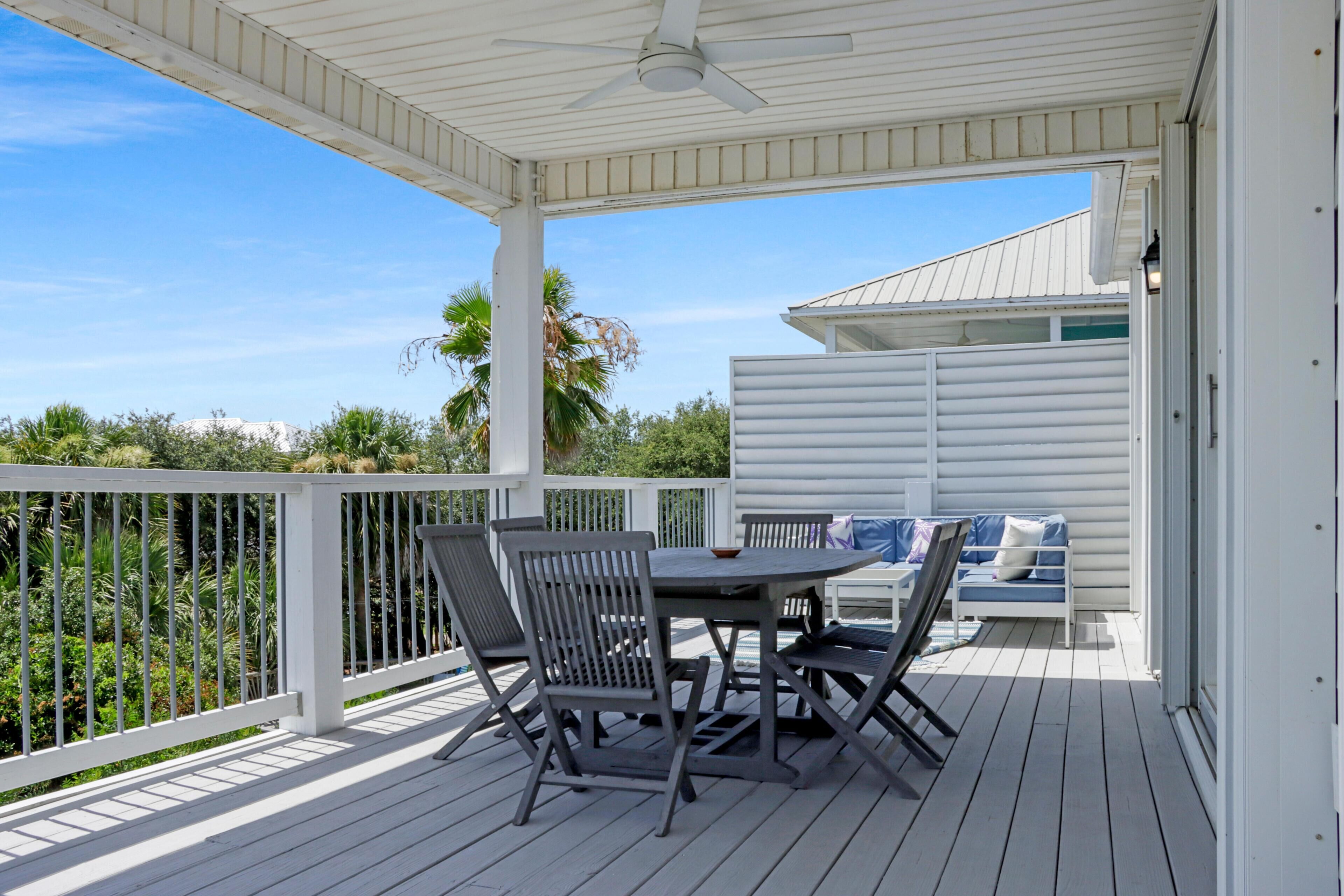 A covered outdoor patio with a table and chairs, a ceiling fan, and a view of the surrounding greenery.