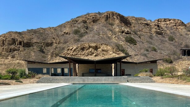 Piscina cristalina con montañas al fondo, un área de estar techada y un edificio con techo plano.