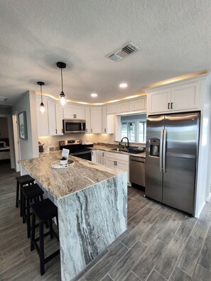 A modern kitchen with white cabinets, a stainless steel refrigerator, and a marble island with bar stools. - Tidewater: Bring your boat, backyard beach,kayaks. (Hernando Beach)