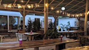 An outdoor seating area with wooden tables and chairs, surrounded by plants and under a covered roof with string lights.