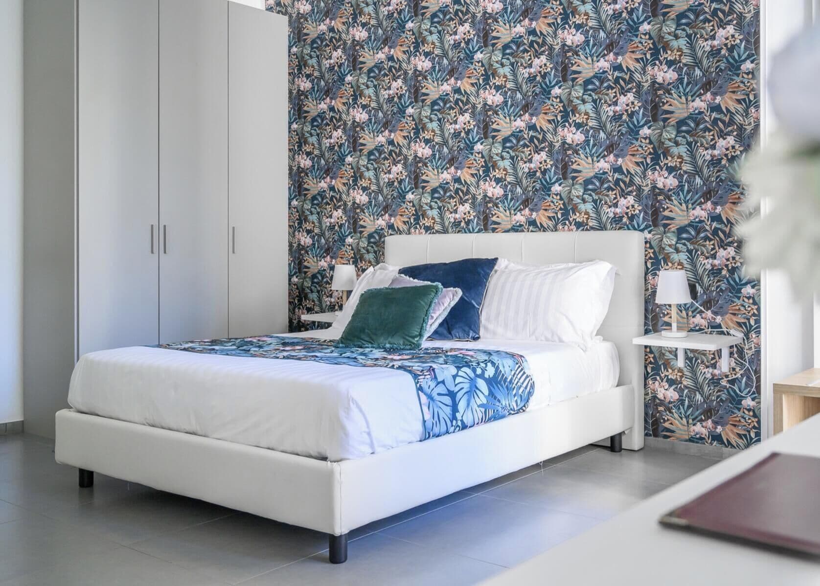 Ein modernes Schlafzimmer mit einem weißen Bett, blumigem Wandpapier und einem Nachttisch mit Lampe.