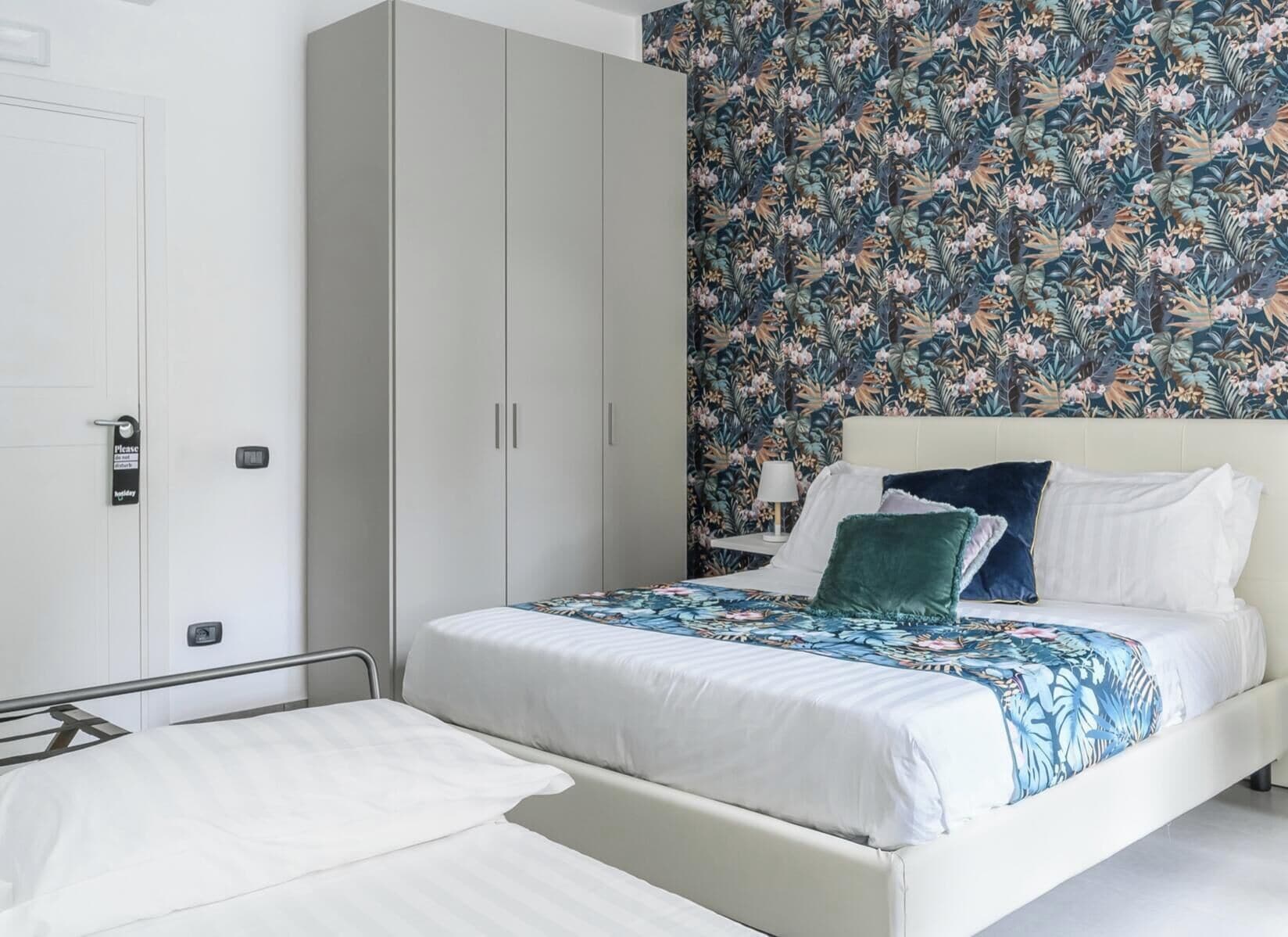 Une chambre d’hôtel avec un lit, une armoire, une table de chevet et un papier peint à motifs floraux.