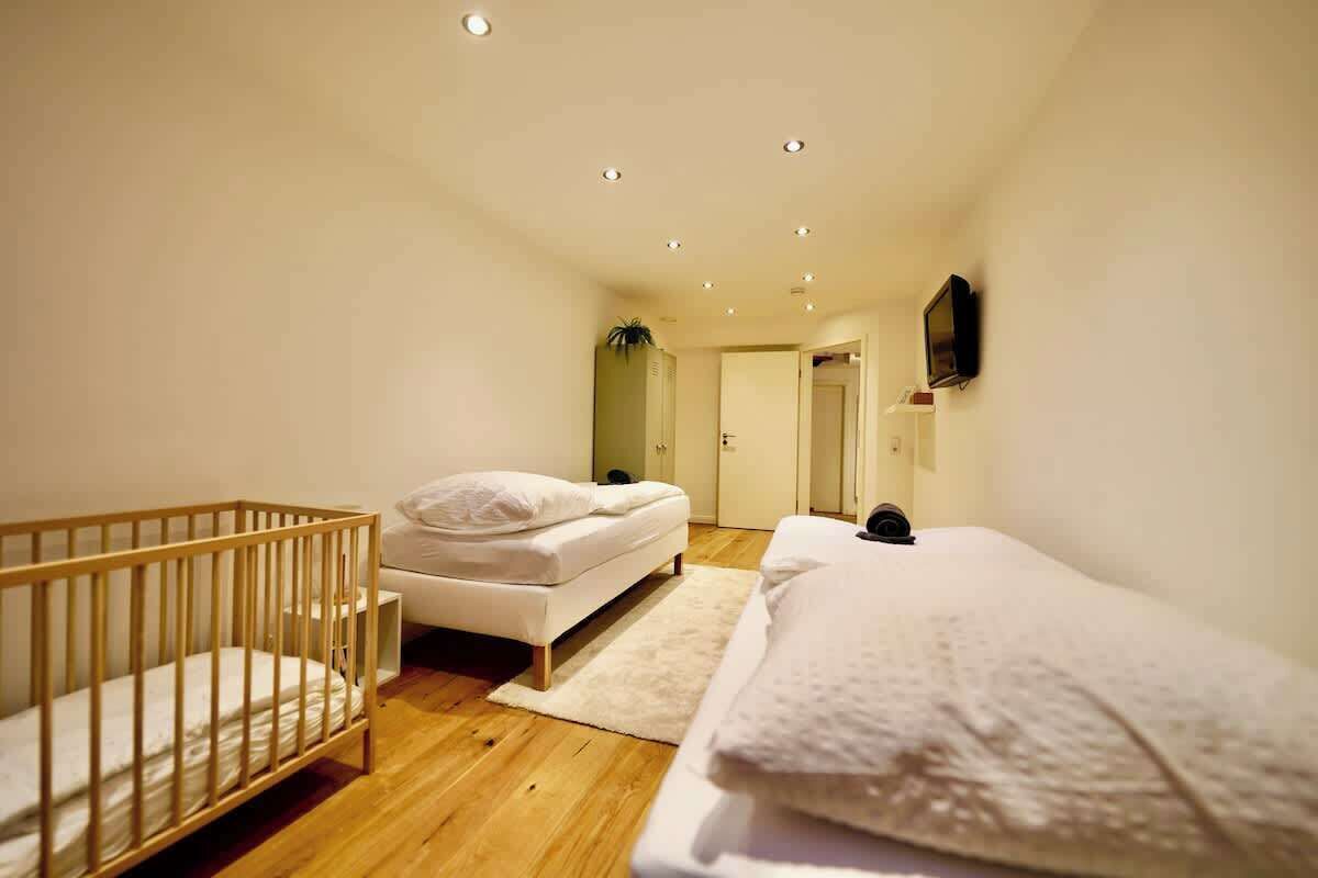 Ein Schlafzimmer mit einem Babybett, einem Bett, einem an der Wand montierten Fernseher und einer Tür zu einem anderen Raum.