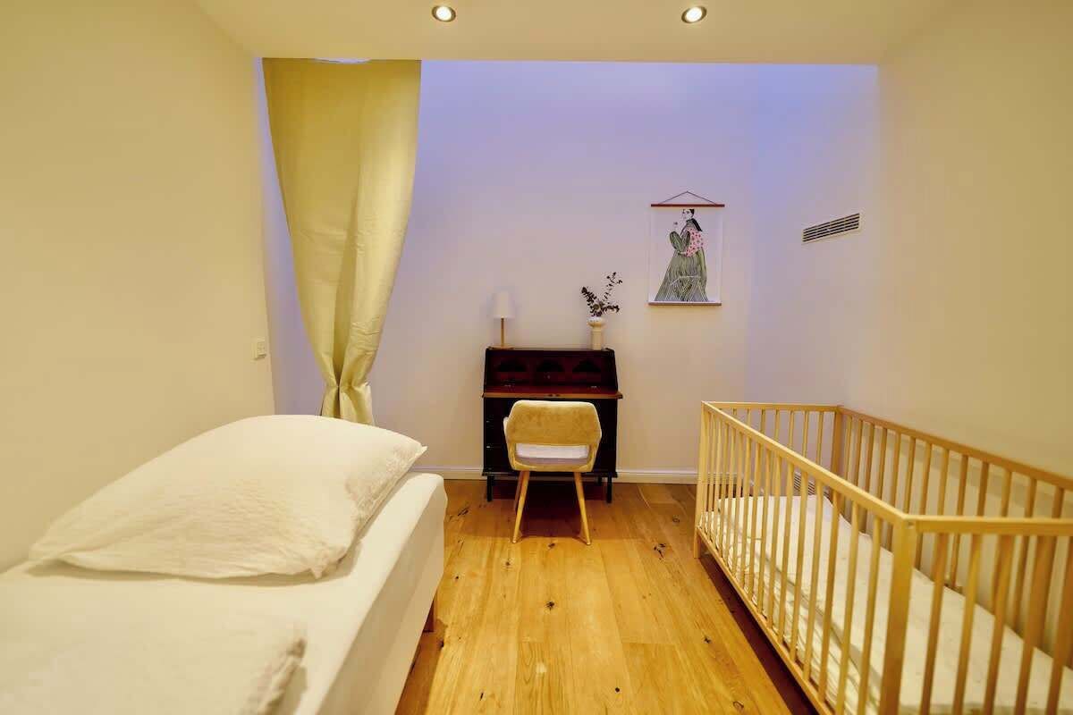 Ein Zimmer mit einem Bett, einem Schreibtisch, einem Sessel, einem Babybett und einem an der Wand hängenden Bild.