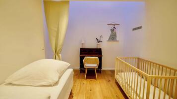 Ein Zimmer mit einem Bett, einem Schreibtisch, einem Sessel, einem Babybett und einem an der Wand hängenden Bild.