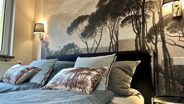 A bedroom with a bed, pillows, a nightstand, a lamp, and a mural of trees on the wall.