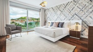 Quarto moderno com uma cama grande, uma mesa lateral, uma cadeira e vista para a cidade através de amplas janelas.