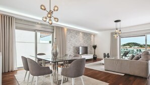 Uma sala de estar moderna com uma mesa de jantar de vidro, cadeiras cinzas, um sofá e uma TV em frente a uma parede de mármore.