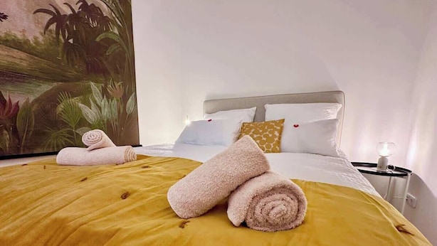 Una cama con toallas enrolladas, una colcha amarilla y una mesita de noche con una lámpara.