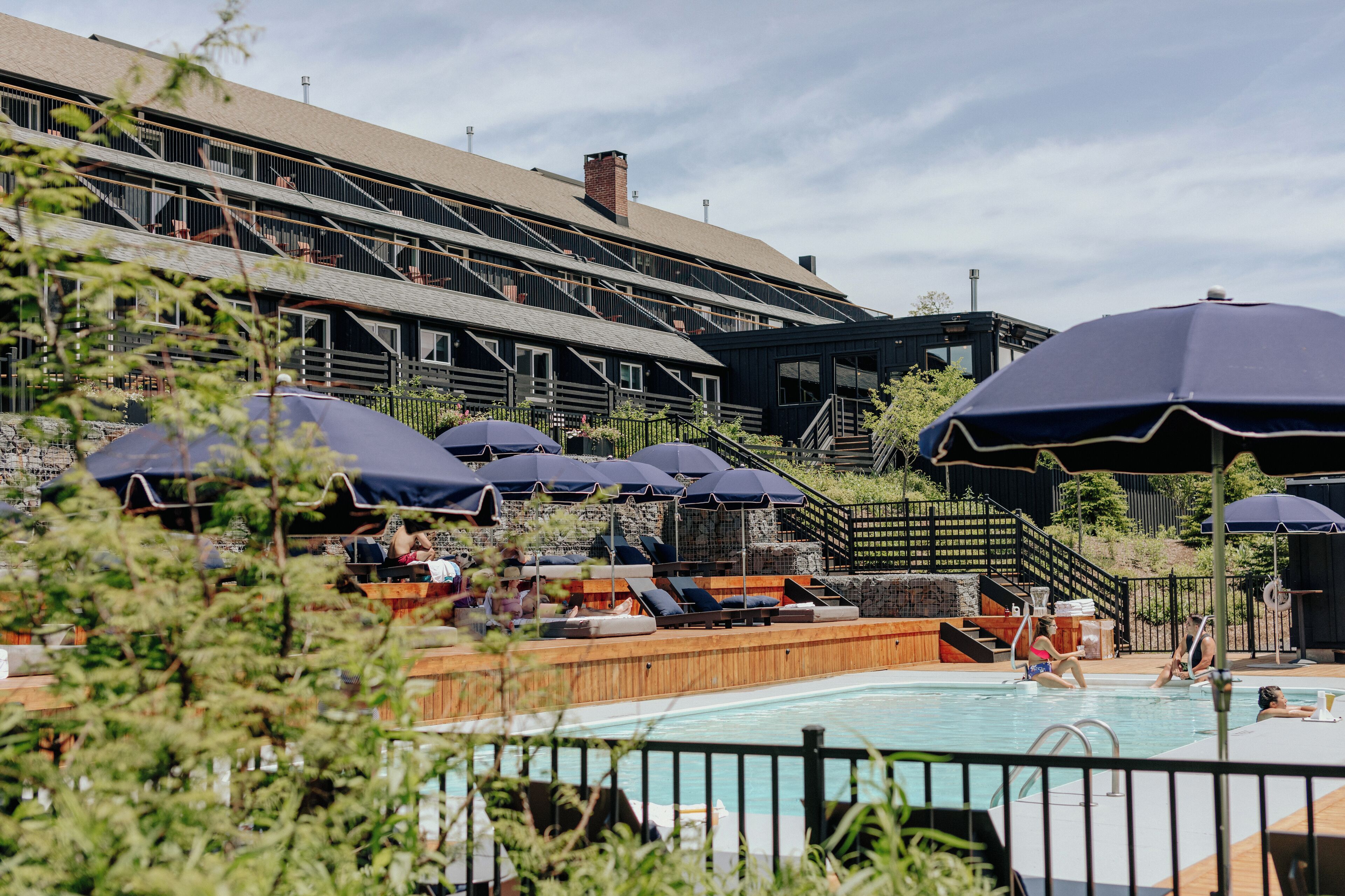 Un espace piscine avec des chaises longues et des parasols, une terrasse en bois et un bâtiment à plusieurs niveaux doté de balcons.