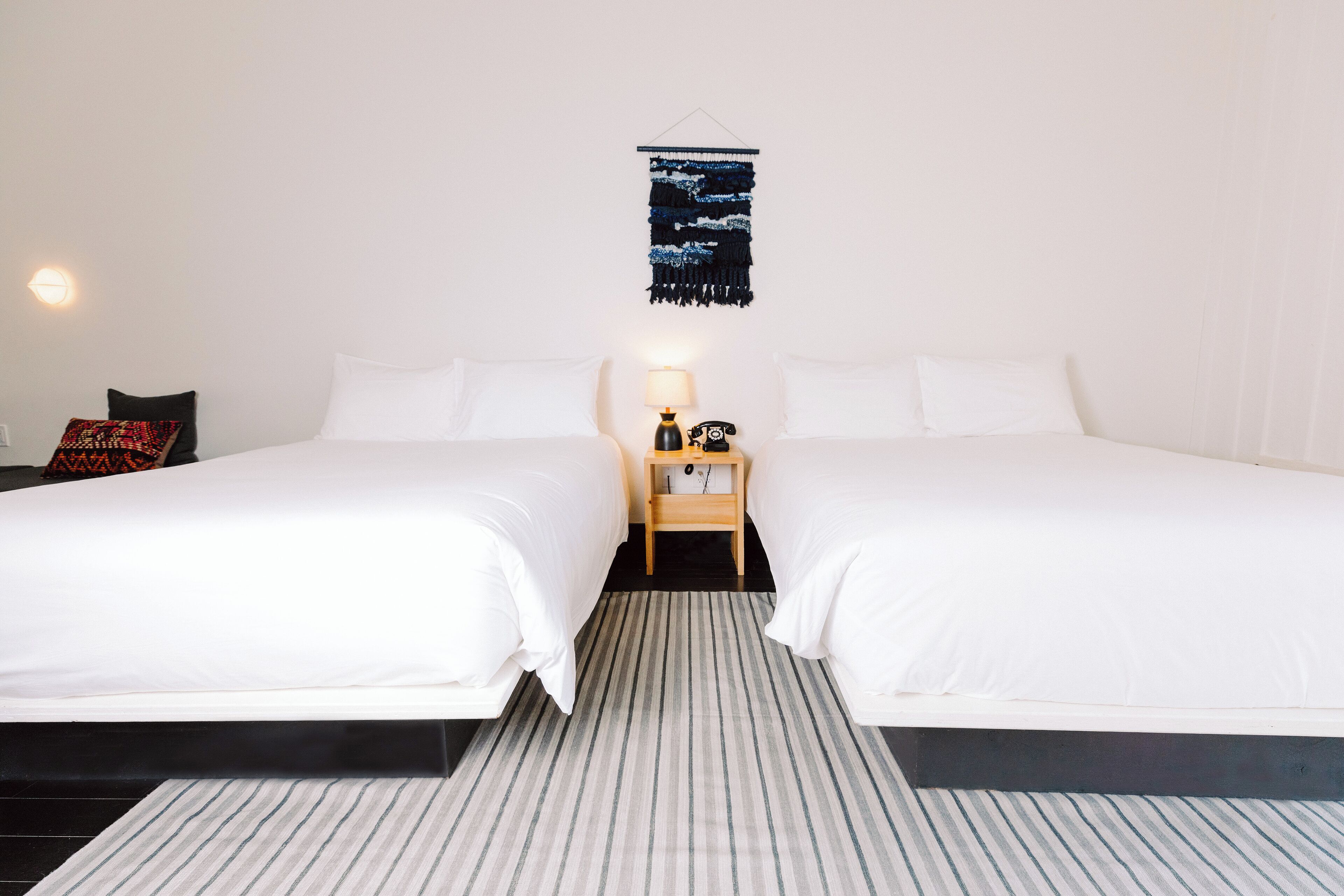 A hotel room with two beds, a striped rug, a wall-mounted tapestry, and a bedside table with a lamp.