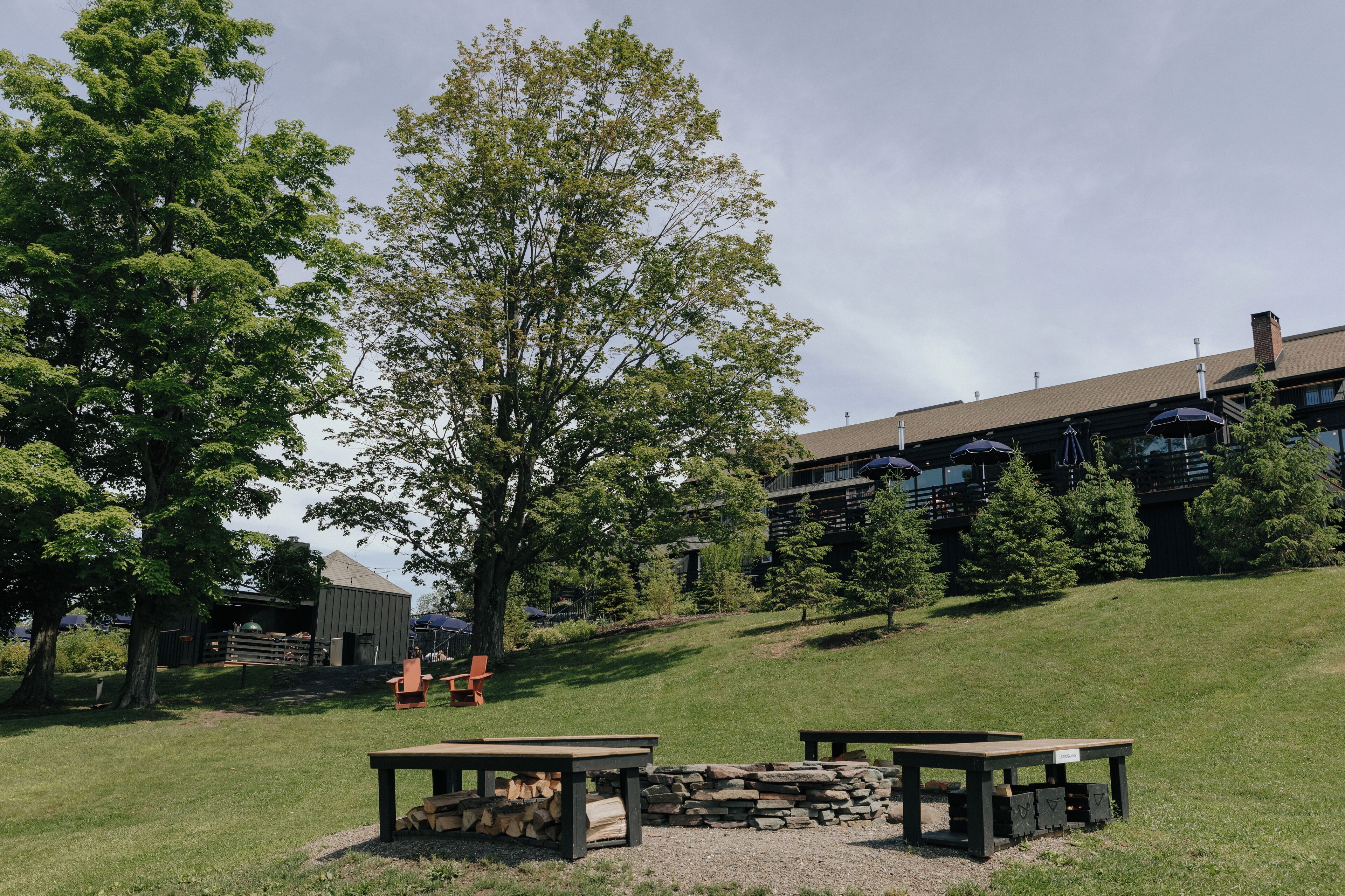 A rustic outdoor seating area with a fire pit, surrounded by a grassy landscape and a large tree.