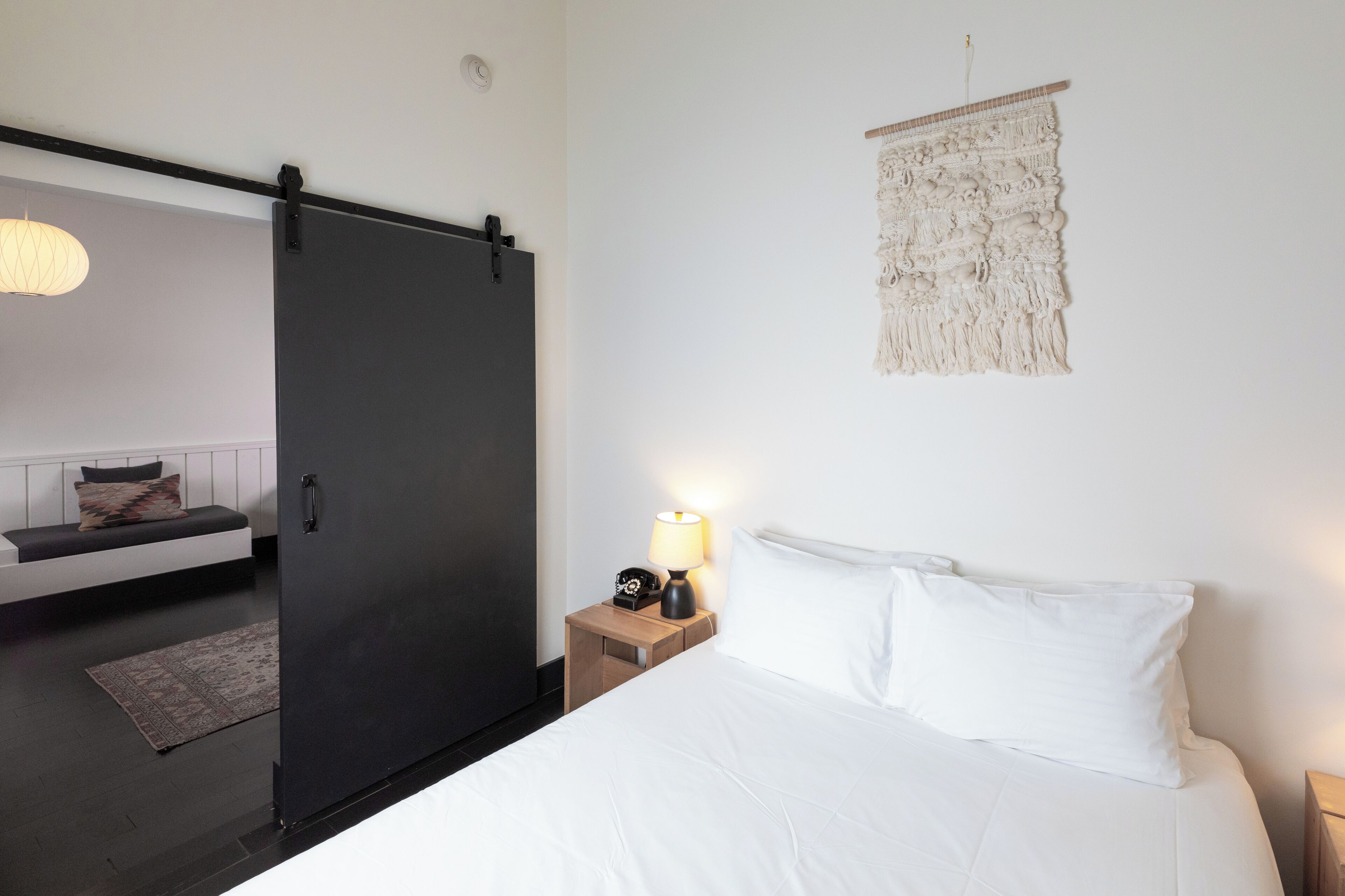 A bedroom with a bed, bedside table with a lamp, a wall hanging, and a sliding door leading to another room.