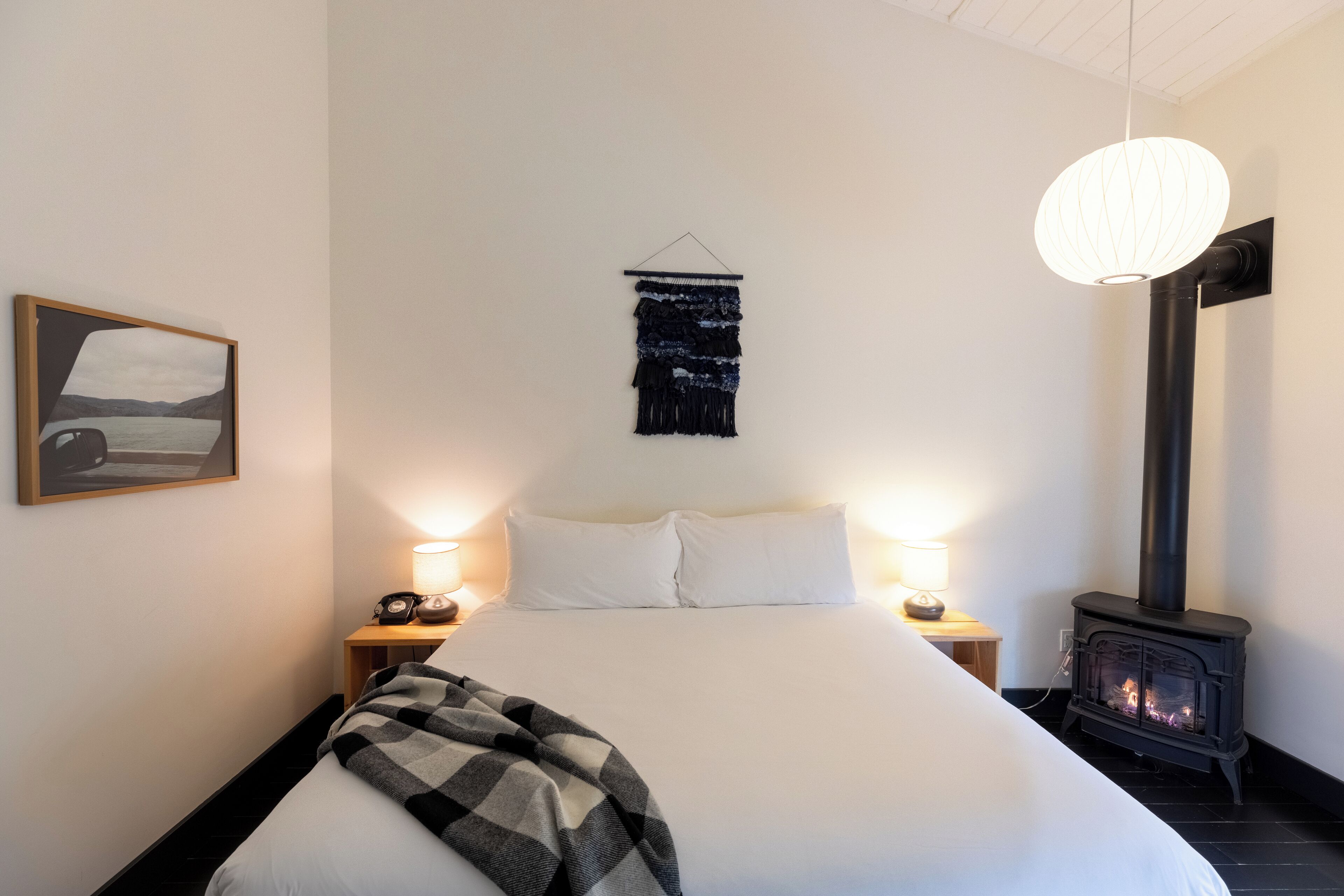 A bedroom with a bed, a wall-mounted tapestry, a hanging light fixture, a nightstand with a lamp, and a wood-burning stove.