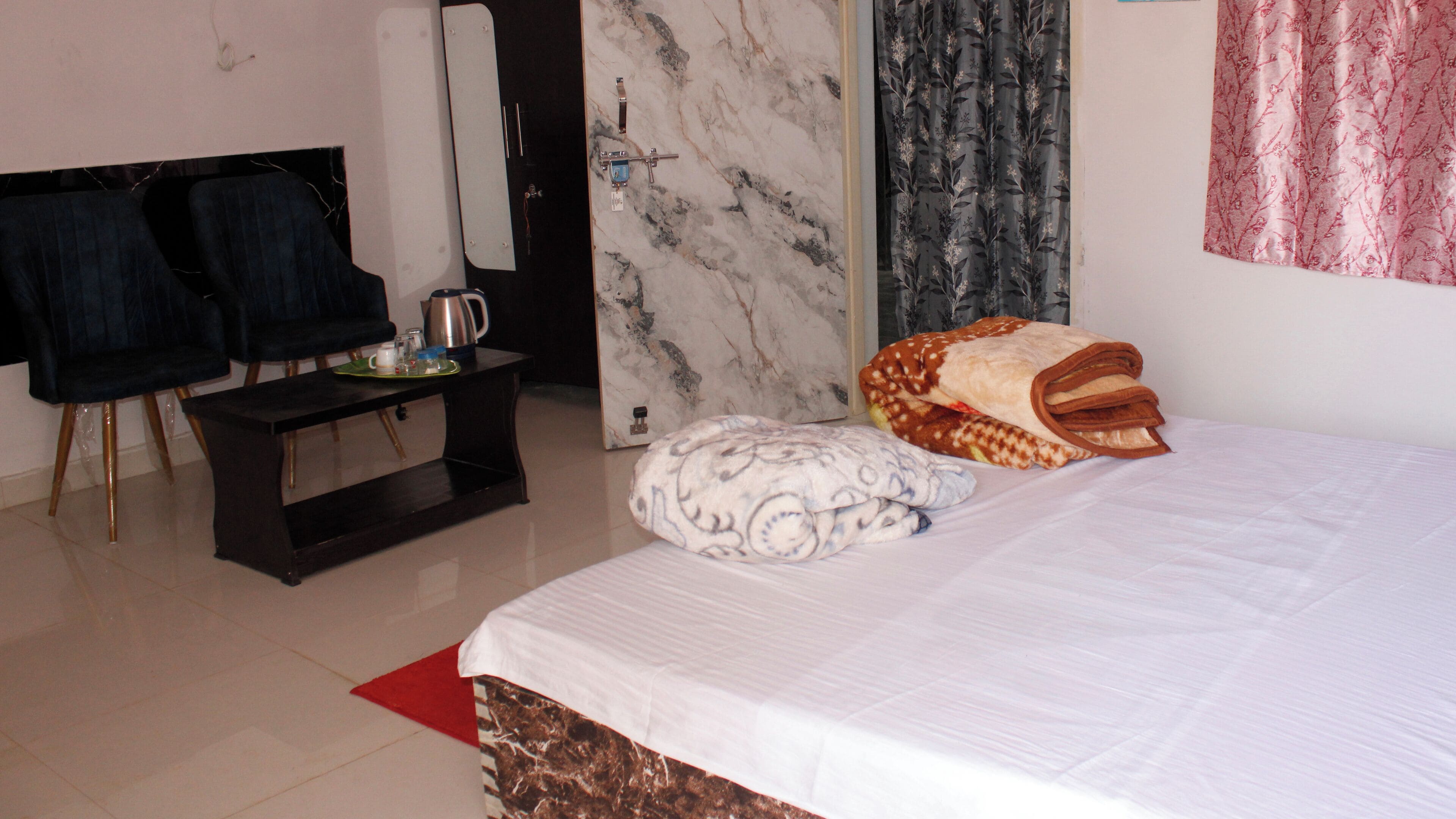 Quarto de hotel com cama, uma pequena mesa com chaleira, cadeiras, espelho e armário.