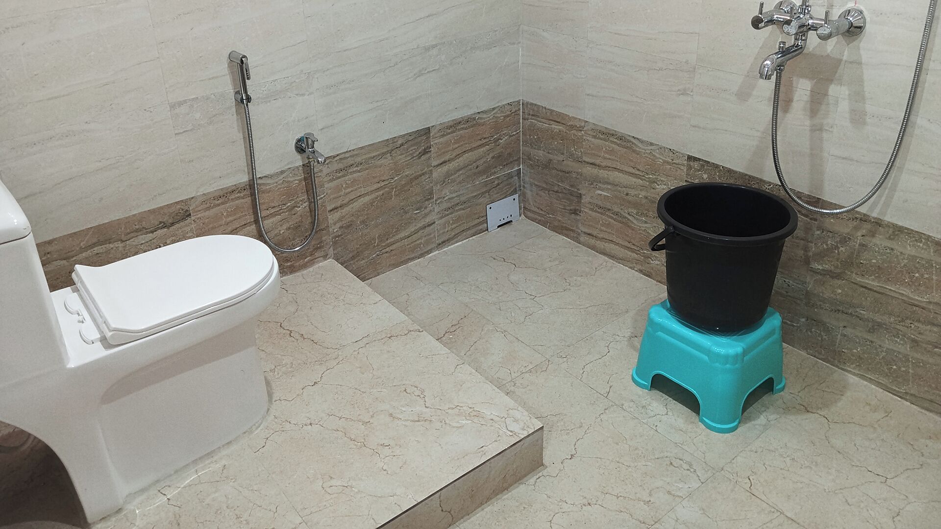 A bathroom with a toilet, a shower, and a black bucket on a turquoise stool.