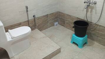 A bathroom with a toilet, a shower, and a black bucket on a turquoise stool.
