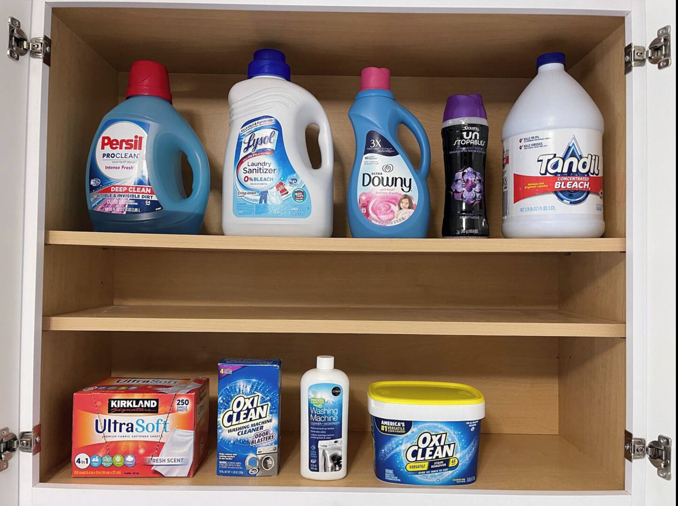 A cabinet with various cleaning products including Persil PROCLEAN, LYSOL Laundry Sanitizer, Downy Ultra, Tandil Concentrated Bleach, Kirkland Signature UltraSoft, OxiClean, and OxiClean Stain Remover.